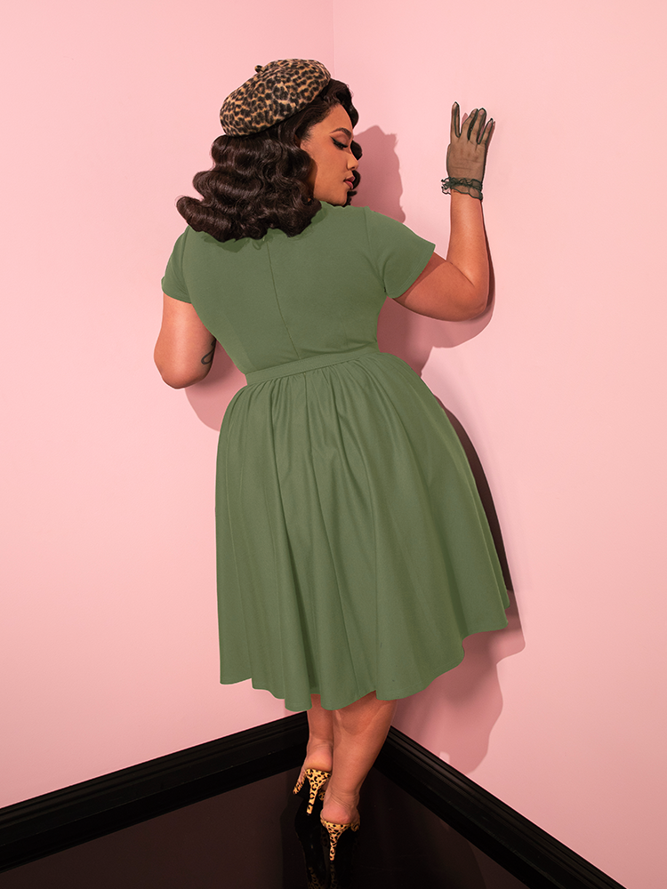 Effortlessly chic, the female model captivates while showcasing the Avon Swing Dress in Sage Green, a stunning creation from the retro brand Vixen Clothing.