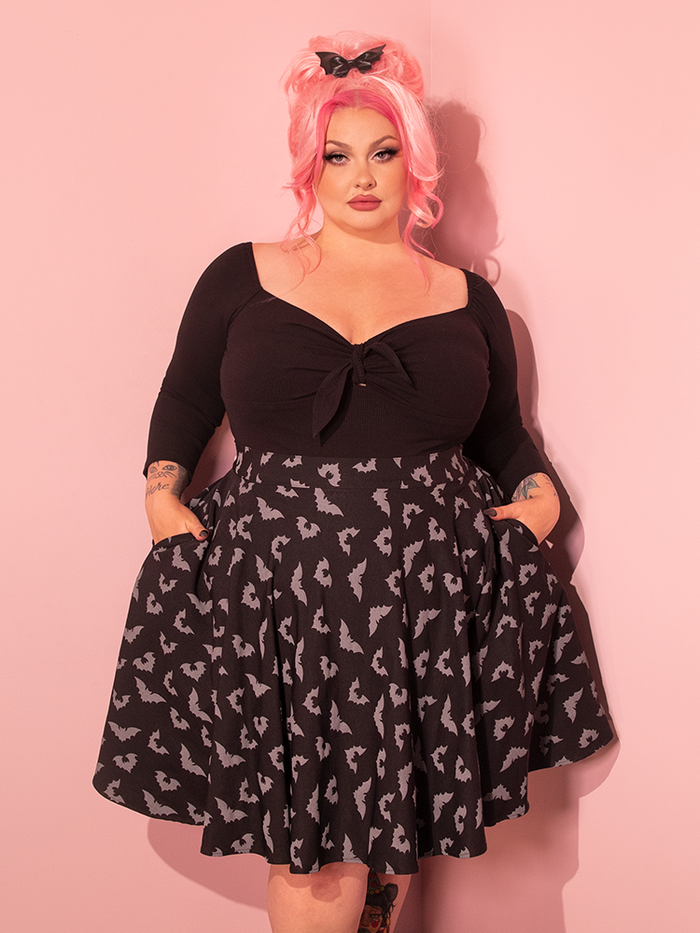 A pin-up fashion icon graces the scene, donning the Maneater Skater Skirt in a captivating Glow-in-the-Dark Bat Print, within a showroom awash with alluring vintage pink.