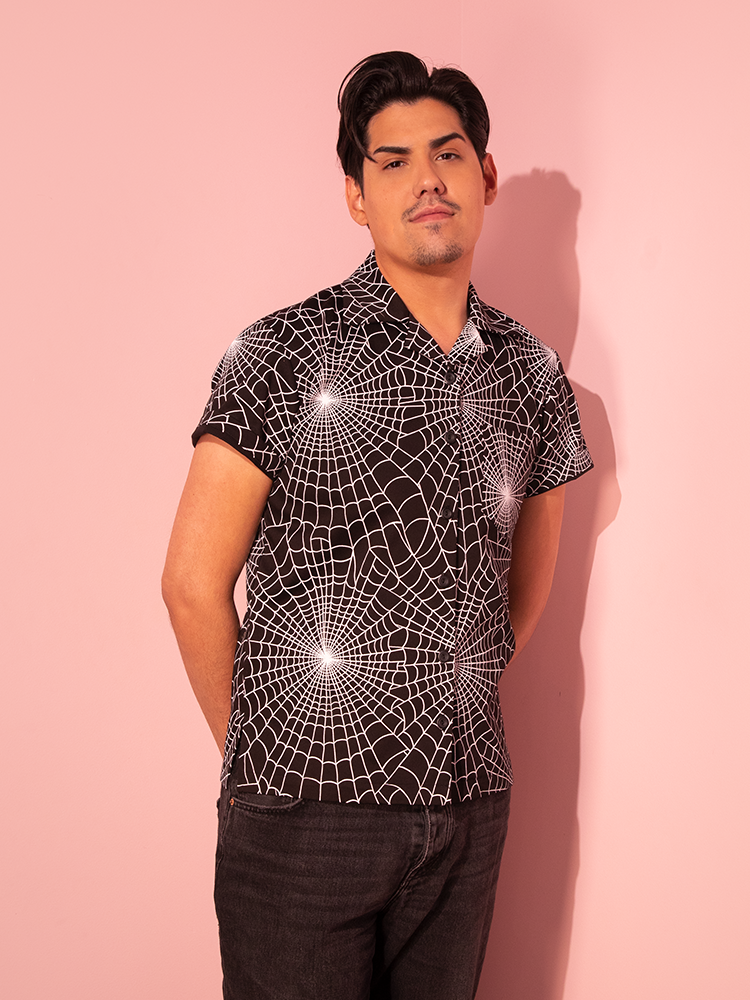 A male model elegantly flaunts the Halloween Spider Web Print Button Up Short Sleeve Shirt in Black, a creation of the retro fashion brand Vixen Clothing.