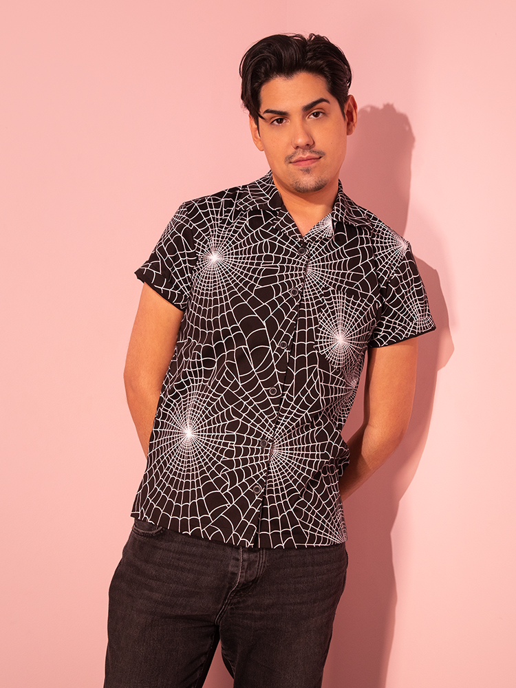 Captured in an arresting shot, a male model dons the Button Up Short Sleeve Shirt in Halloween Spider Web Print (Black) from the nostalgic Vixen Clothing line.