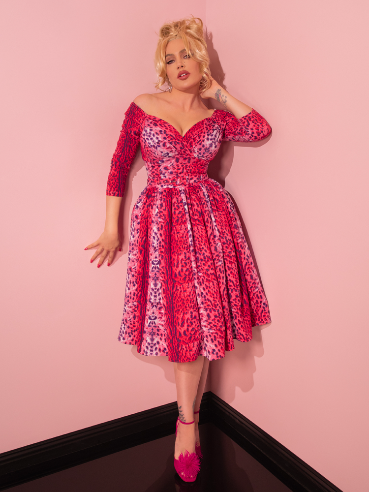 Embodying the essence of vintage sophistication, a seductive model captivates with her alluring pose while highlighting the Pink Leopard Print Starlet Swing Dress and Scarf, a timeless creation by Vixen Clothing, a beloved retro dress and clothing retailer.