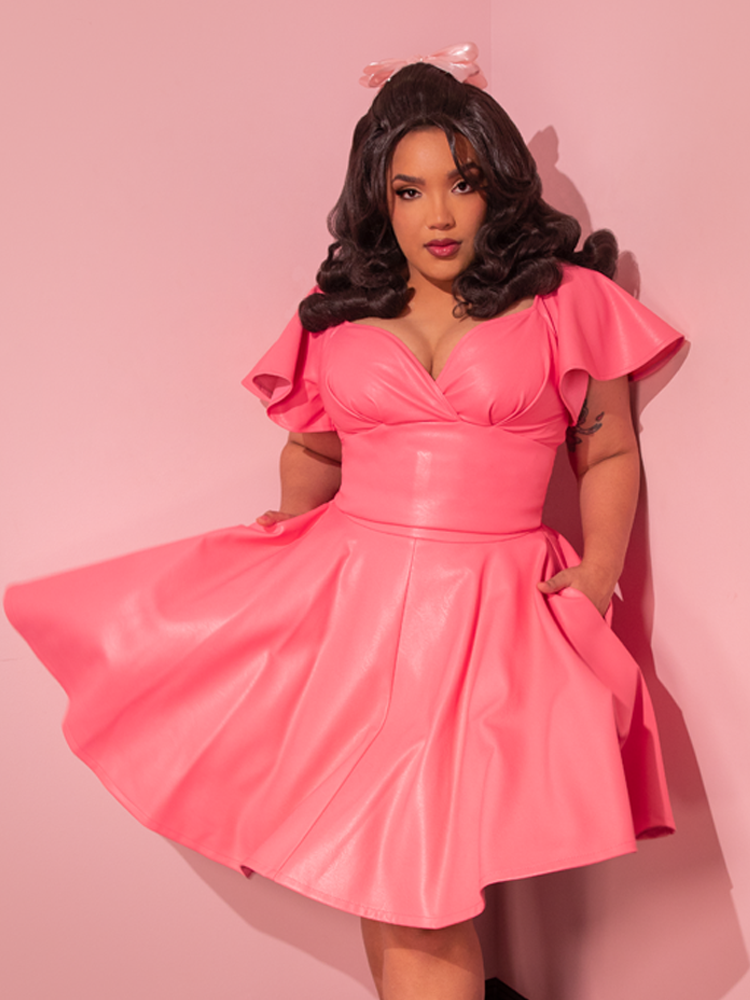 Embracing the retro charm, the stunning model confidently flaunts the Flamingo Pink Vegan Leather Bad Girl Skater Skirt, a vintage-inspired gem crafted by the esteemed retro dress maker Vixen Clothing.