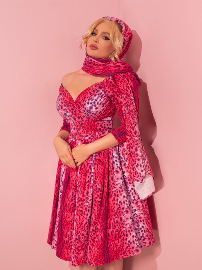 Embracing the allure of retro fashion, a seductive model confidently showcases the Pink Leopard Print Starlet Swing Dress and Scarf from Vixen Clothing, a renowned vintage-inspired dress and clothing retailer.