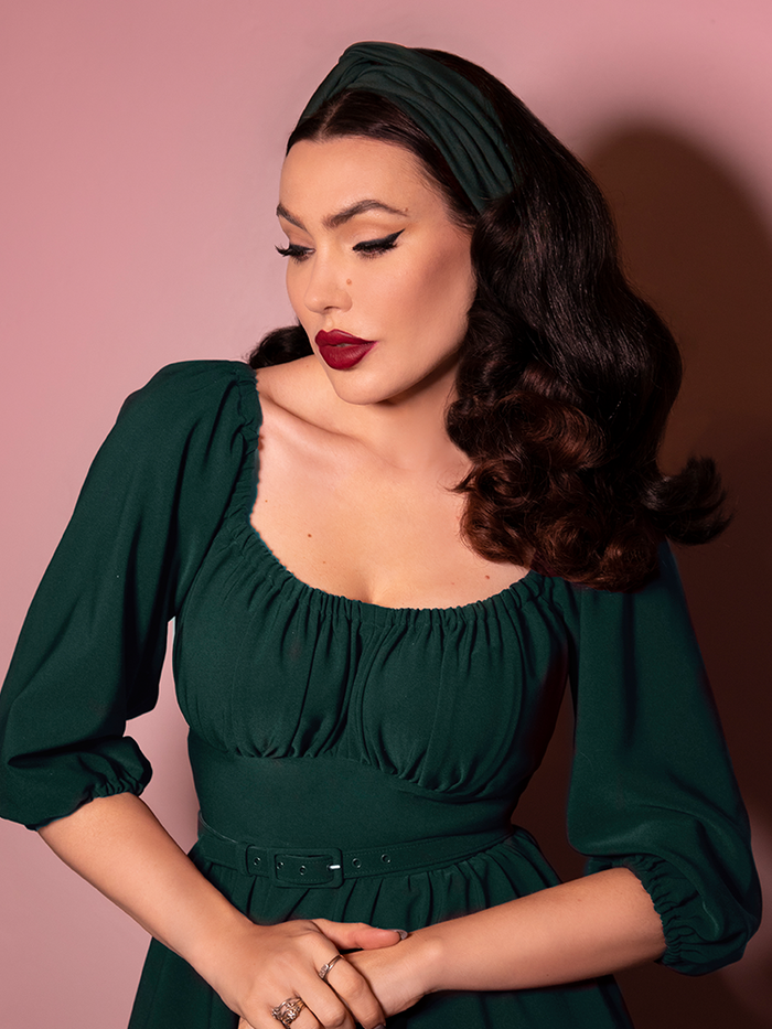 A closeup of Micheline Pitt looking down modeling the vintage style knot headband in hunter green paired with a matching dress.