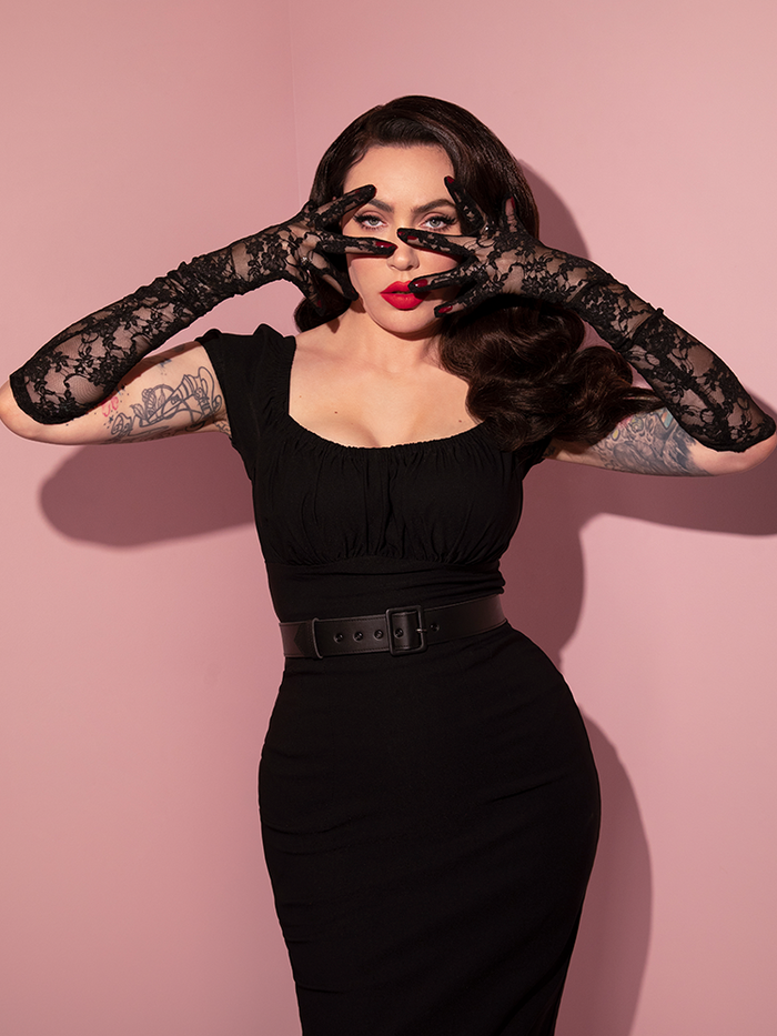 Micheline Pitt poses with her arms and hands, slightly covering her face, draped in the Floral Lace Gloves in Black.