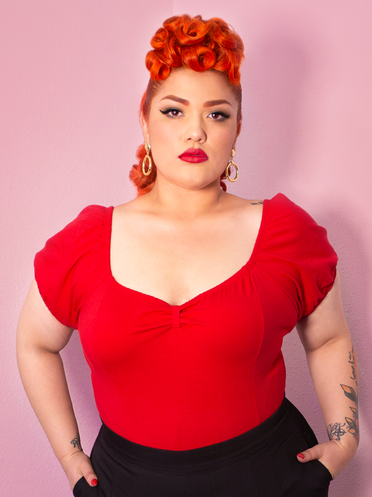 Evelyn looking at the camera with her hands in her pockets modeling the Powder Puff top in red by Vixen Clothing.