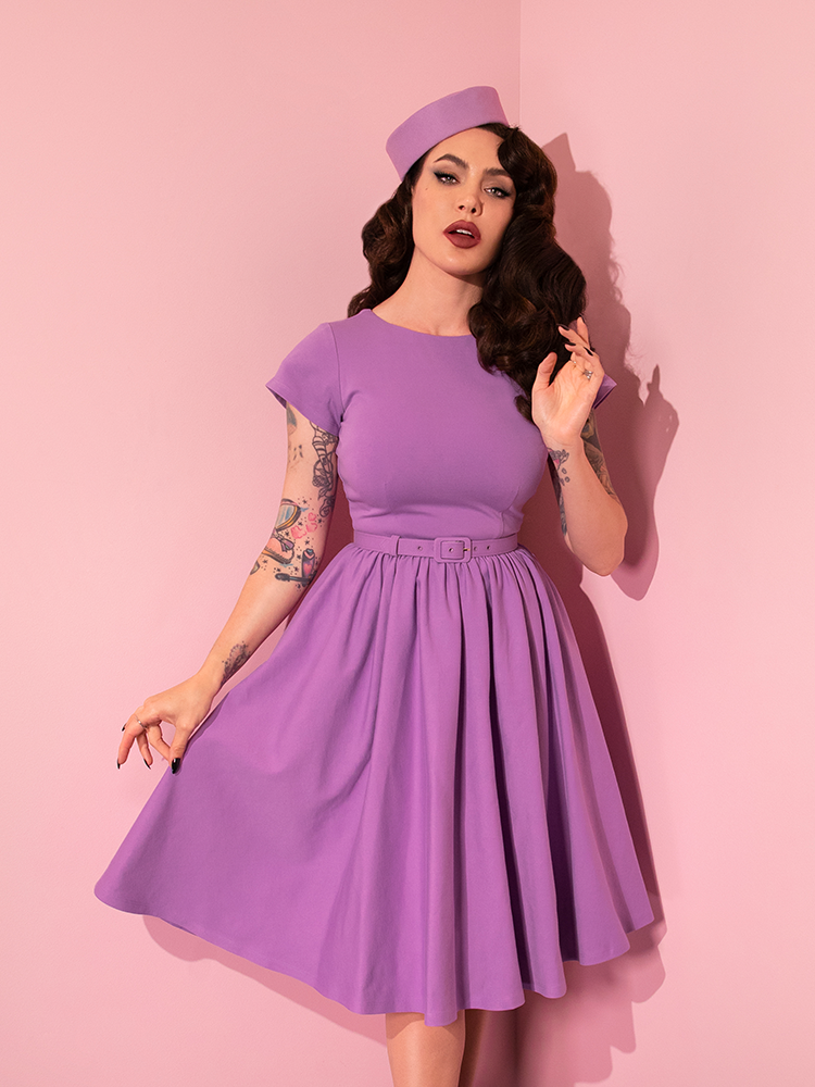 Micheline Pitt standing and posing with one hand held close to her face and the other gently pulling out the skirt section of the Avon Swing Dress in Lilac.