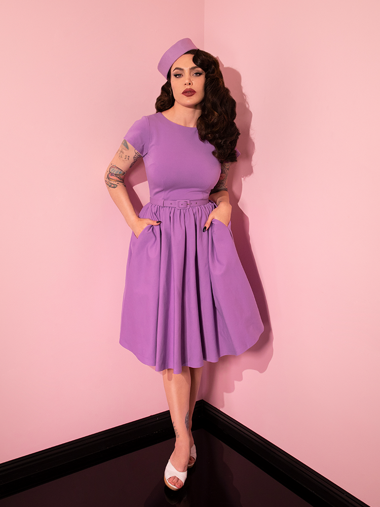 The Avon Swing Dress in Lilac worn by Micheline Pitt who is tucking her hands into the side pockets.