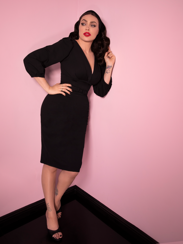 Leaning against the wall, Micheline Pitt looks off to the side while modeling the Bawdy Wiggle Dress in Black from Vixen Clothing.