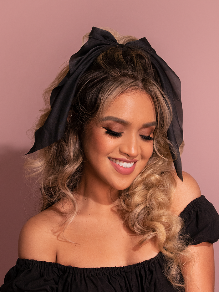 The female retro model showcases the vintage-inspired Large Satin Hair Bow in Black from the iconic retro brand Vixen Clothing.