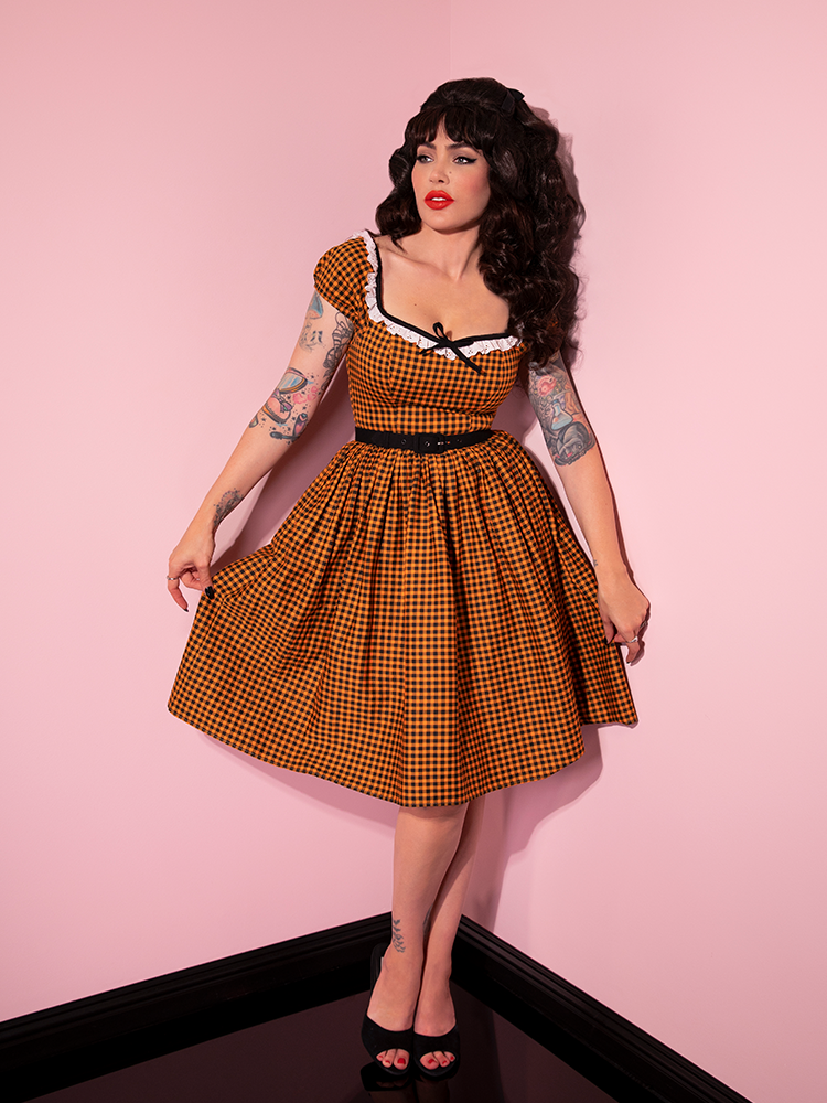 Micheline Pitt looking off camera and pulling on the sides of the skirt on her Bardot Beauty Swing Dress in Orange Pumpkin Gingham.