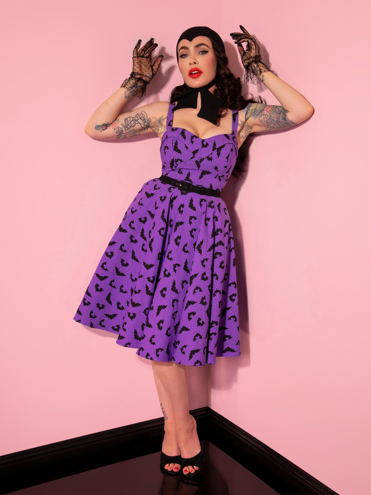Micheline Pitt, wearing the Maneater Swing Dress in Bat Print, poses with hands held up by her head.