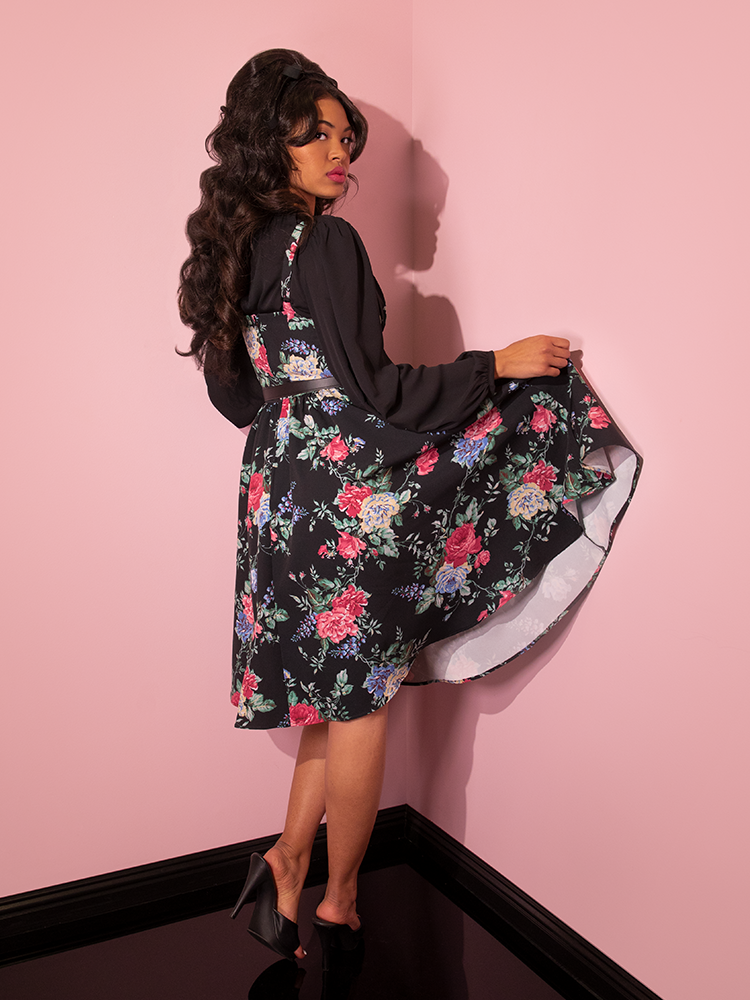 Danelly turned away from the camera and slightly pulls up the skirt section of the Ingenue Dress in Black Rose Print.