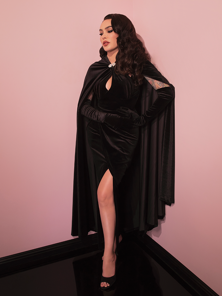 Micheline Pitt pensively looking down while showing off the Golden Era Cape in Black Velvet from retro dress brand Vixen Clothing.