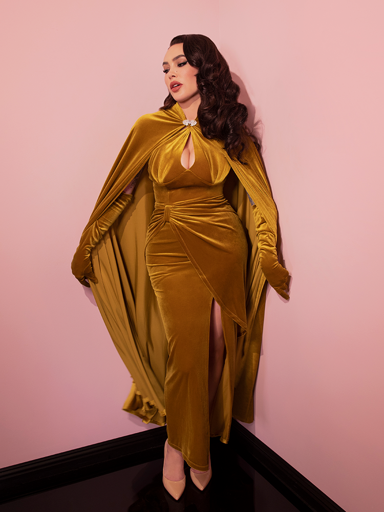 Micheline Pitt pulling out the sides of the Golden Era Cape in Gold Velvet along with her matching elbow length gloves, retro style dress and heels.