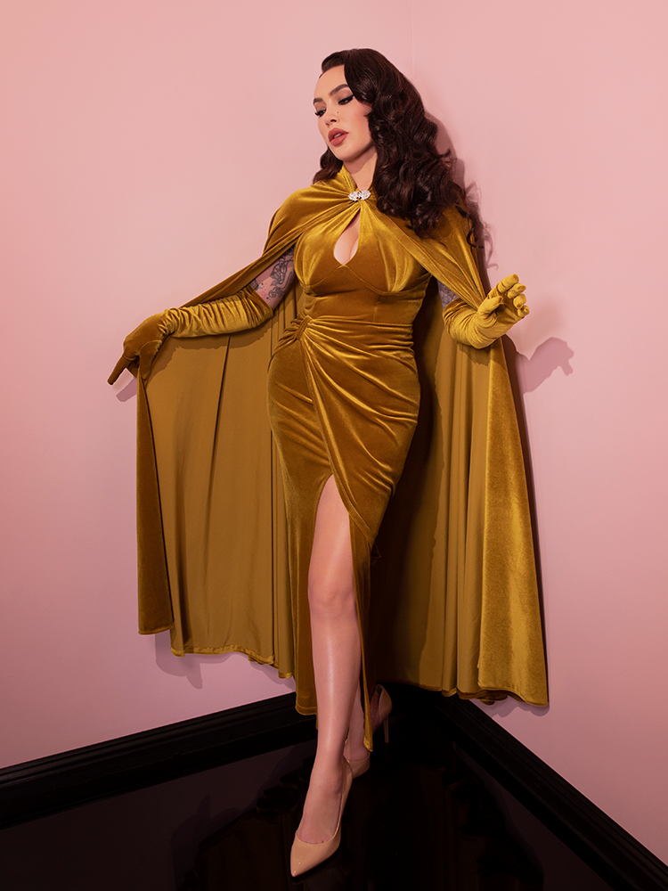 Micheline Pitt posing with her arms outstretched by her side while wearing the Golden Era Cape in Gold Velvet over the matching dress and elbow length gloves. All items from Vixen Clothing