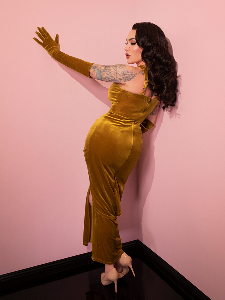 Micheline is facing and leaning against a pink wall while wearing the Golden Era gown and glove set.