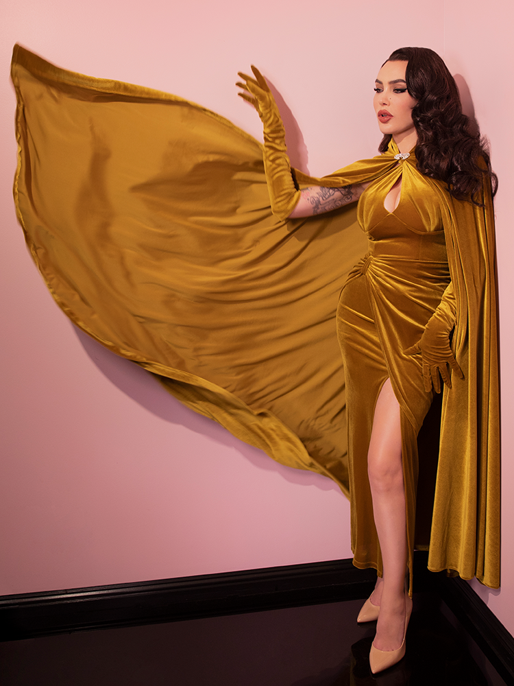 Micheline Pitt standing in a pink showroom, tosses out the side of the  Golden Era Cape in Gold Velvet to show off it’s dramatic length.