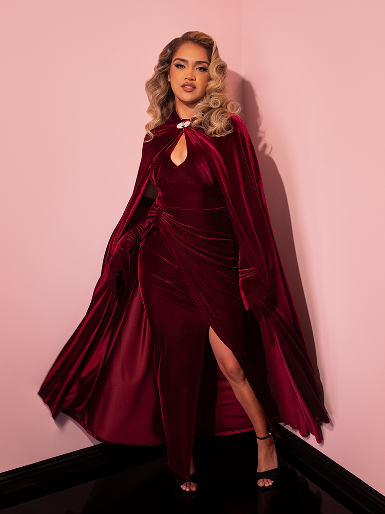 Model posing and looking directly into the camera while wearing a burgundy velvet gown and the matching Golden Era Cape in Burgundy Velvet.