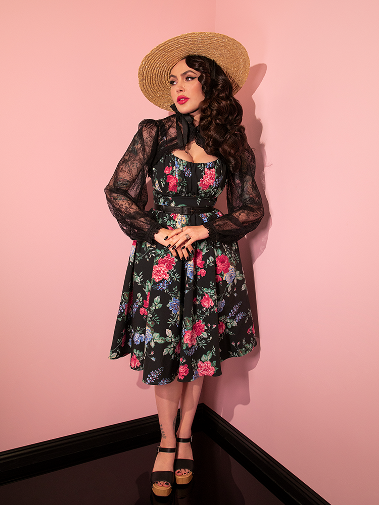 Micheline Pitt with her hands clasped at her waist, wears the Ingenue Dress in Black Rose Print.