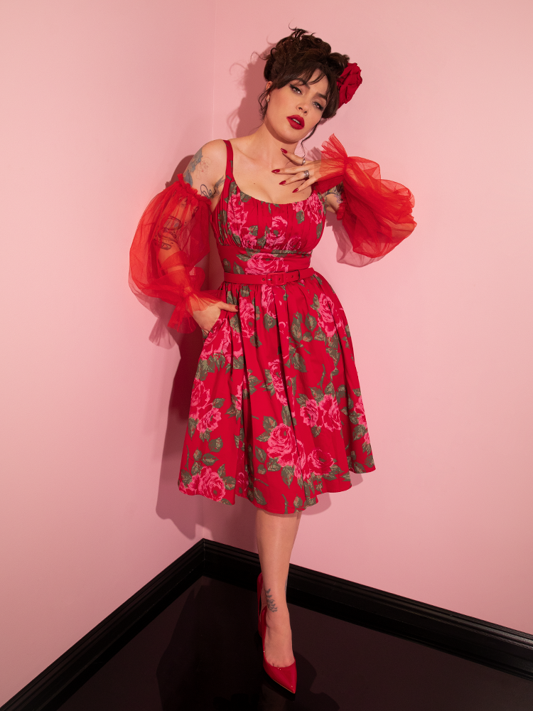 Micheline Pitt tucks her hand into the pocket of her Ingenue Swing Dress in Vintage Red Rose Print while wearing the red tulle sleeves - also from retro clothing company Vixen Clothing.