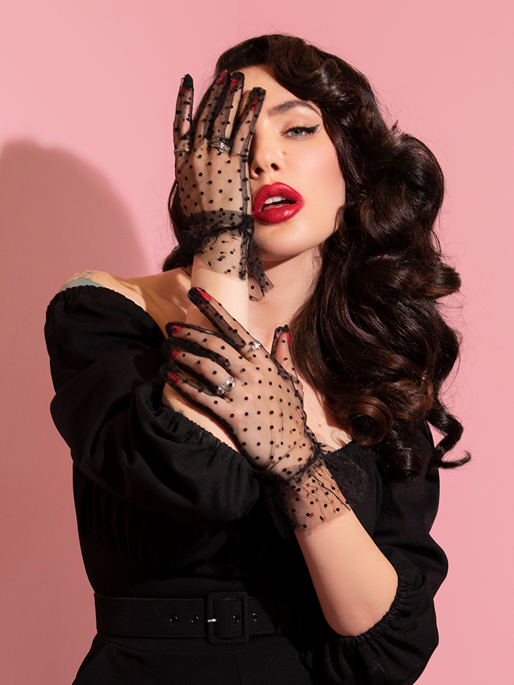 Covering half of her face with her hand that is covered in the Mesh Polka Dot Gloves in Black, Micheline Pitt looks directly into the camera.