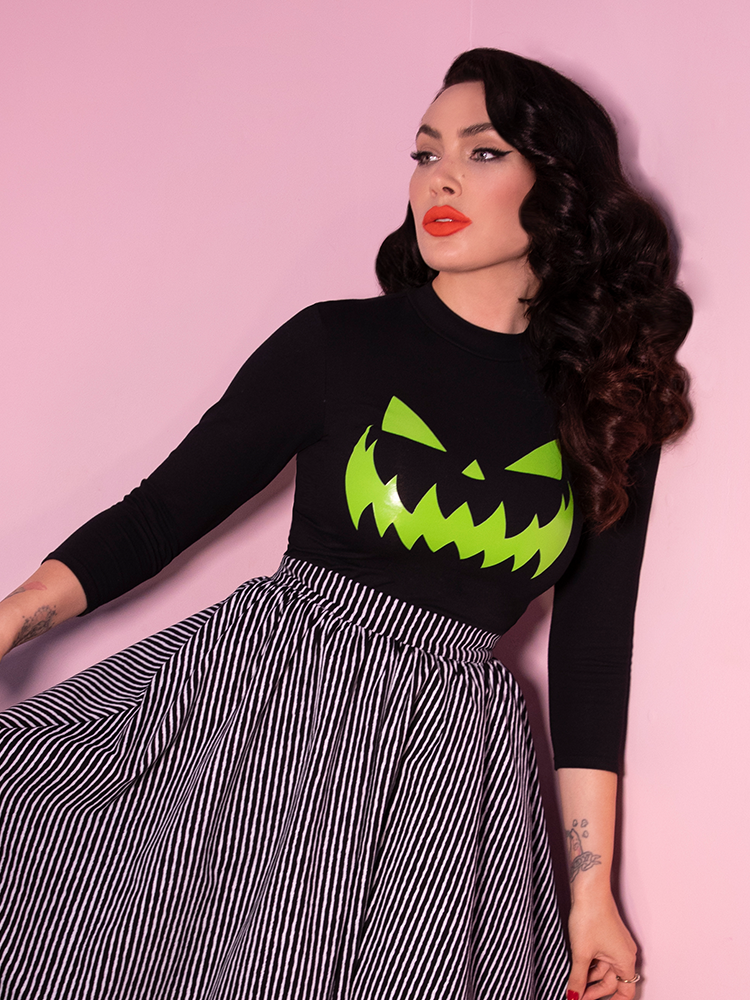 Looking off camera and tugging at the sides of her black and white striped skirt, Micheline Pitt wears the Black Pumpkin King Glow in the Dark 3/4 length sleeve retro top.