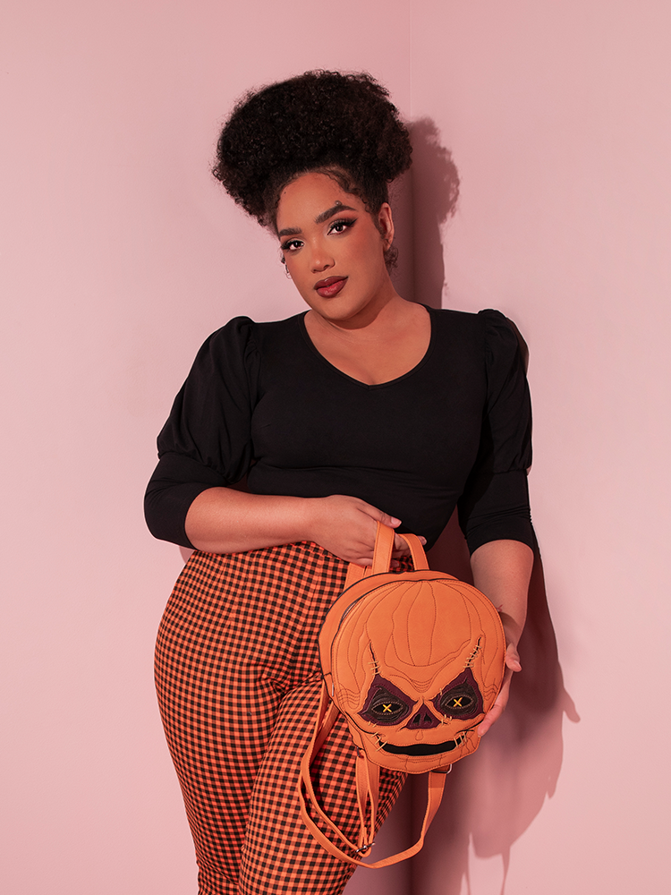 Female model leading against the wall while holding the TRICK R TREAT™ Sam Backpack with a retro style outfit including black retro top and gingham pants.