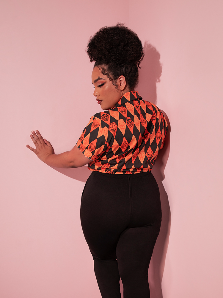 Female model turned away from the camera to show off the back of the TRICK R TREAT™ Button Up Shirt in Halloween Harlequin Print which she's tucked into her black cigarette pants to pull off the ultimate retro clothing outfit.