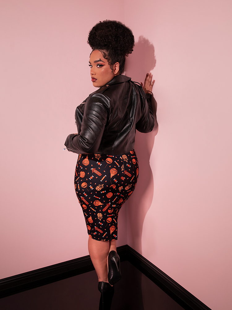 Model turned away from the camera shows off the back of the TRICK R TREAT™ Peplum Wiggle Dress in Candy Corn Novelty Print with a leather jacket pulled over.