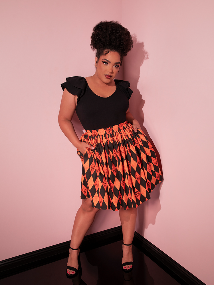 Model tucking her hands into the side pockets of the TRICK R TREAT™ Skater Skirt in Halloween Harlequin Print she's paired with a black retro top.