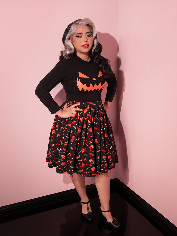Model posing with her hands on her hips and slightly turned to the side while wearing the TRICK R TREAT™ Skater Skirt in Candy Corn Novelty Print with a long sleeved black pumpkin shirt tucked in.