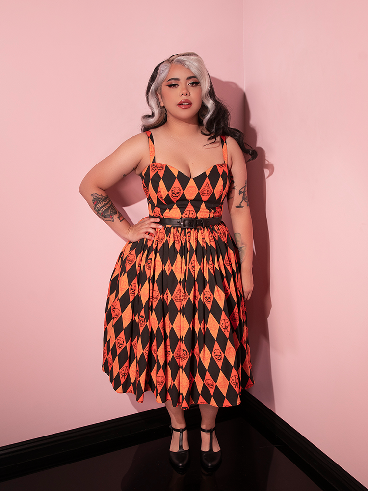 Model posing with one hand resting on her hip and the other hanging down by her hip wearing the TRICK R TREAT™ Sweetheart Swing Dress in Halloween Harlequin Print from retro dress brand Vixen Clothing.