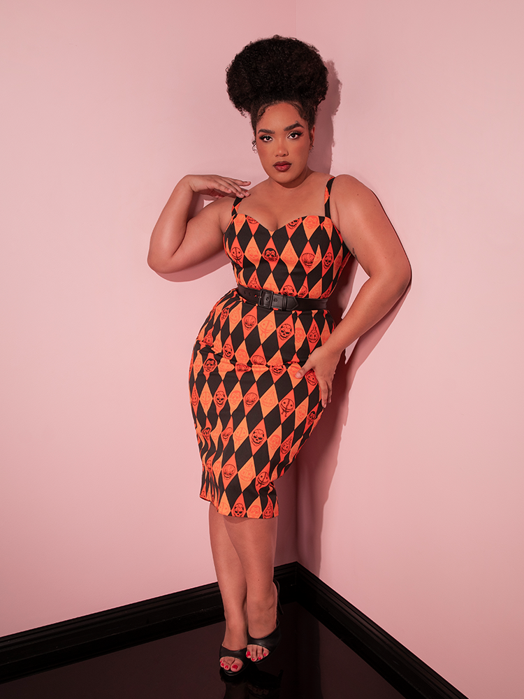 Model leaning back against a pink wall while wearing the TRICK R TREAT™ Sweetheart Wiggle Dress in Halloween Harlequin Print from Vixen Clothing.