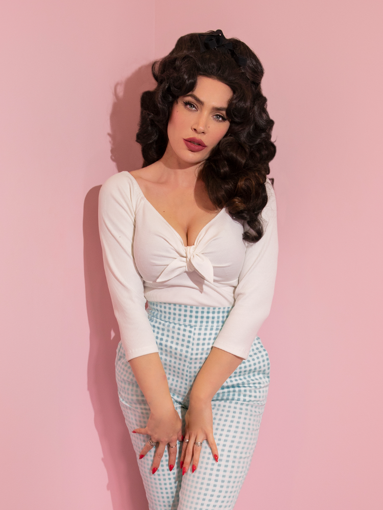 A closeup of Micheline Pitt looking at the camera modeling the tie me up top in white by Vixen Clothing paired with blue gingham pants.