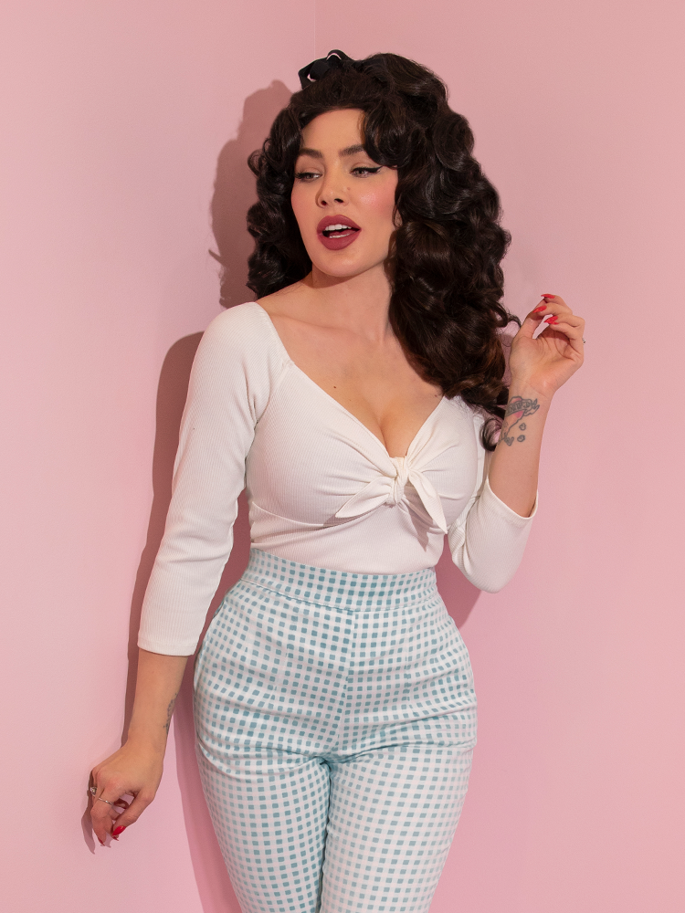 A closeup of Micheline Pitt looking away from camera modeling the tie me up top in white by Vixen Clothing paired with blue gingham pants.