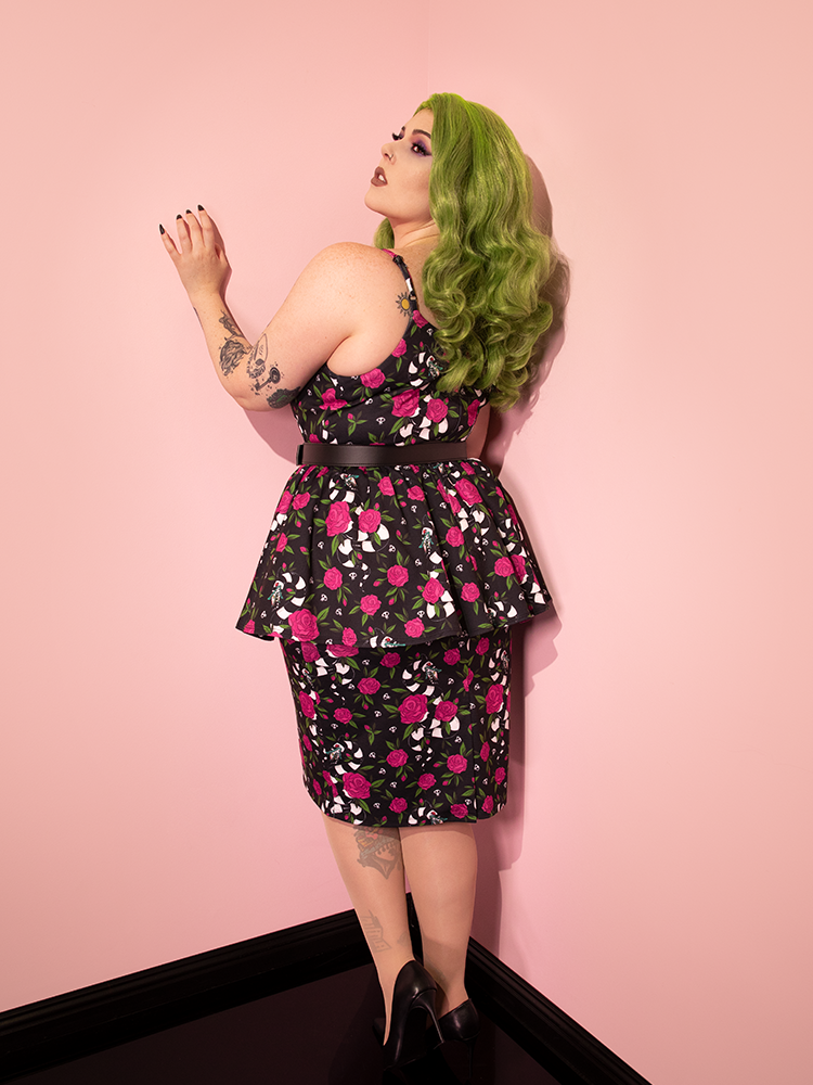 Green haired model facing the wall to show off the back of the BEETLEJUICE™ Sandworm & Roses Peplum Wiggle Dress from Vixen Clothing.