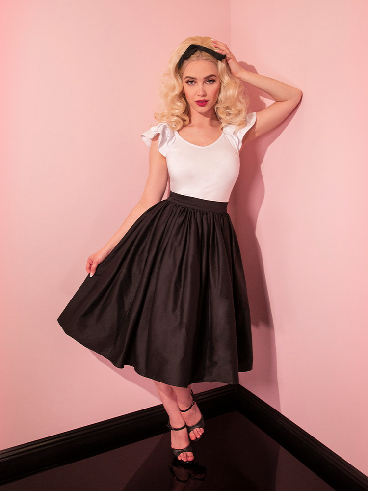 Blonde model posing in the corner of a room with the Vixen Swing Skirt in Black and a white short sleeve top.