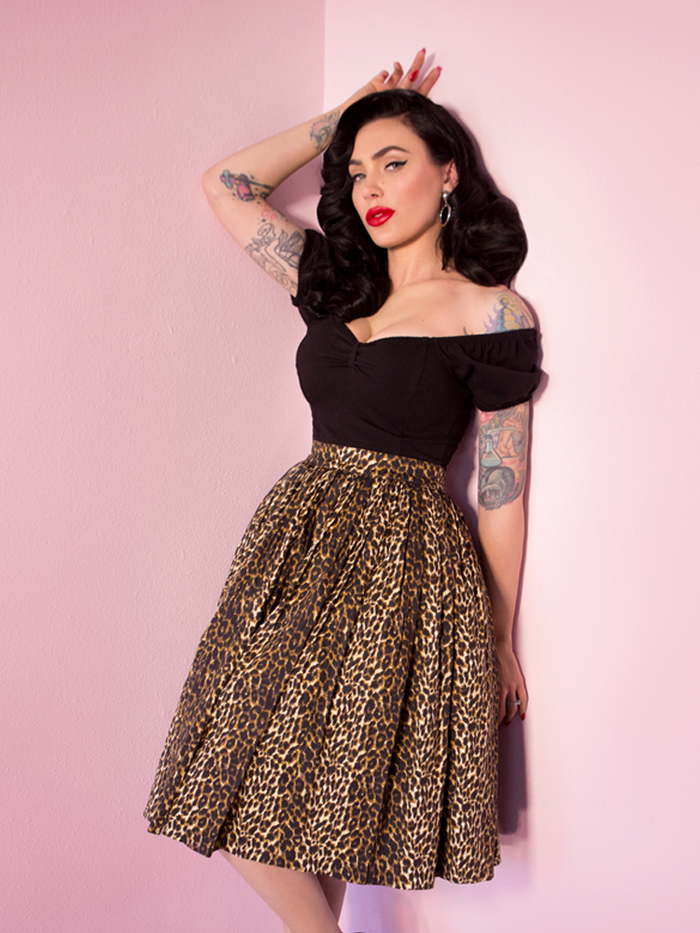 Micheline Pitt leans against the pink walls of her studio while modeling the Vintage Swing Skirt in Wild Leopard Print along with a black off the shoulder top.