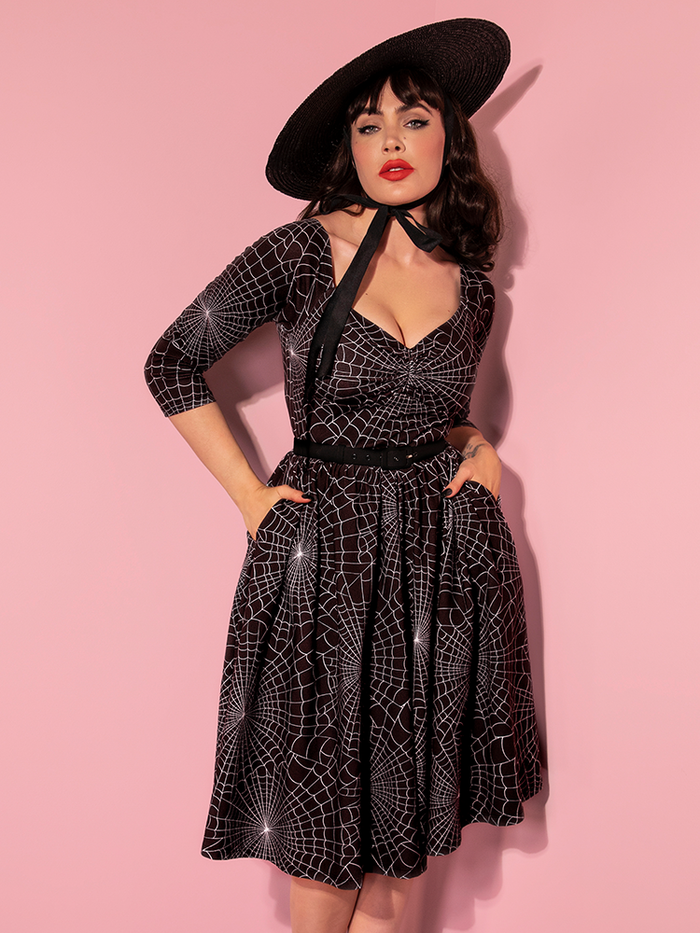 Micheline Pitt posing with her hands in her pockets while wearing the Wicked Swing Dress in Vintage Spiderweb Print.