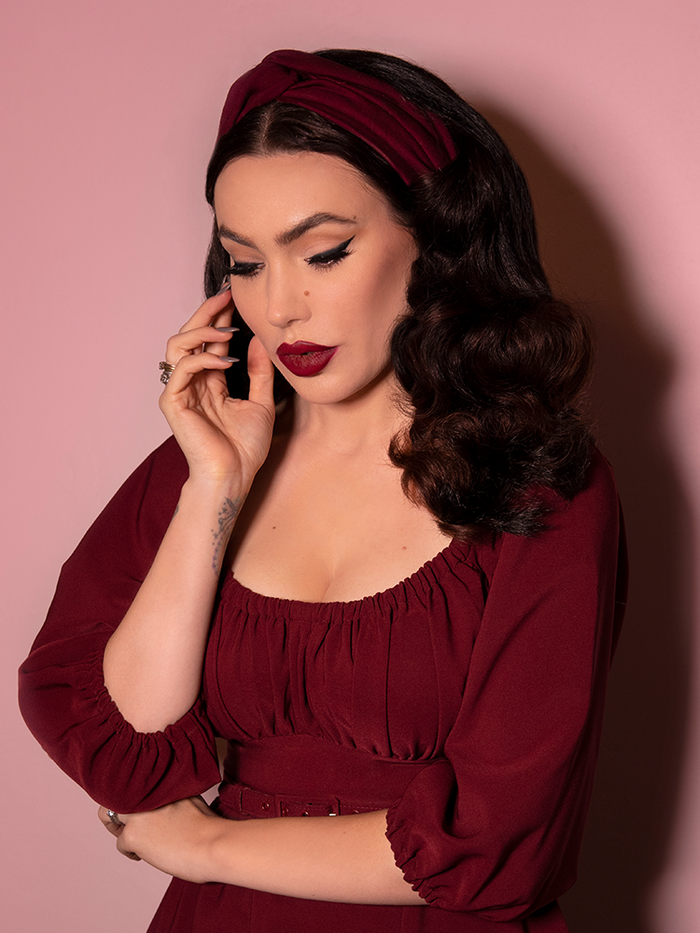 A closeup shot of Micheline Pitt looking down while modeling the knot headband in burgundy paired with a matching dress.
