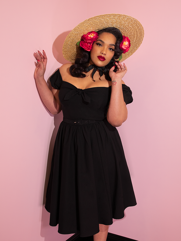 A closeup of Ashleeta wearing a straw hat and flowers in her hair modeling the Vixen dress in black.