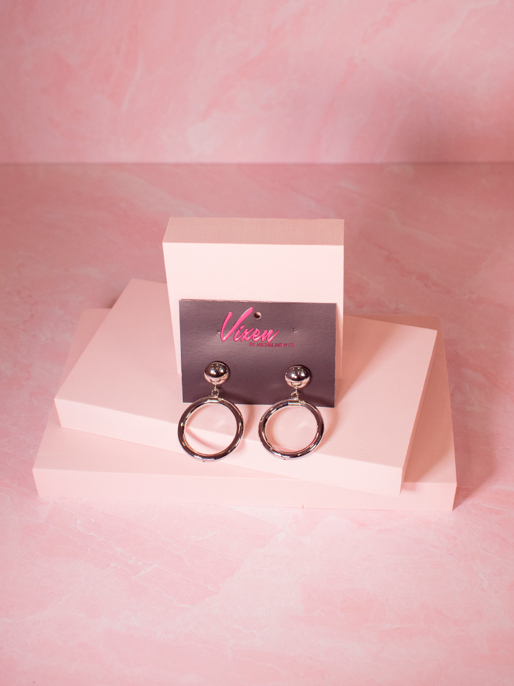 The Bad Girl Hoop Earrings in Silver In original packaging photographed sitting on a pink background.