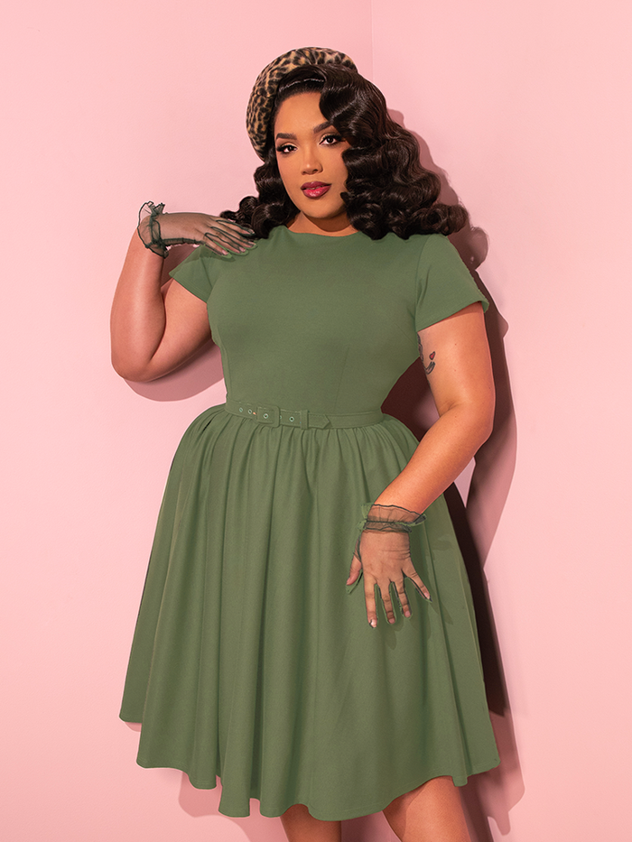 The stunning female model elegantly poses while showcasing the Avon Swing Dress in Sage Green from the retro fashion label Vixen Clothing.