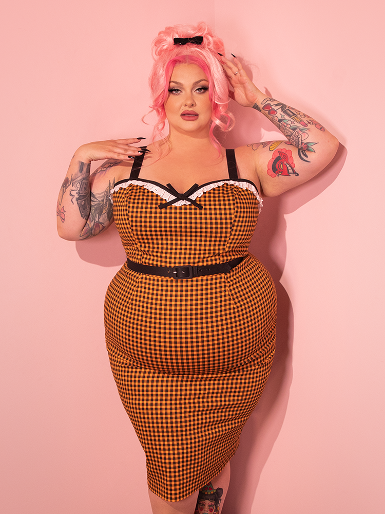 Let the allure of our retro-era model captivate you as she dons the Bardot Beauty Wiggle Dress, featuring Orange Gingham, designed by Vixen Clothing, the premier vintage clothing and retro dress brand.