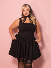A vintage inspired black mini bombshell swing dress delivers classic charm with a flirty twist, styled against a pink wall.