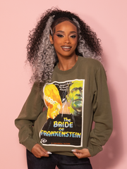 A green sweatshirt featuring a bold Bride of Frankenstein™ vintage movie poster graphic, set against a pink wall.