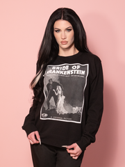A black sweatshirt featuring a bold vintage movie advertisement of the Bride of Frankenstein™, set against a pink wall.