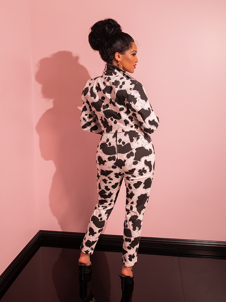 With a vintage flair, the female model joyfully sings and strikes a pose in the all-pink showroom, captivating attention in the Cow Print Cigarette Pants from retro clothing brand Vixen Clothing.