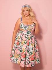 A vintage style cottage corset dress in white garden floral print stands sweetly against a pink wall, evoking pastoral romance.